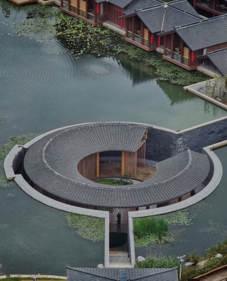 An aerial view of a circular grey tiled roof set on a pond, with traditional buildings behind.