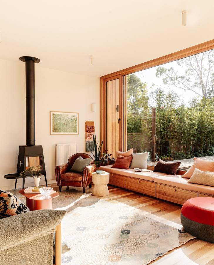 A living room with a long timber window seat under a picture window, and a black wood heater on the wall.