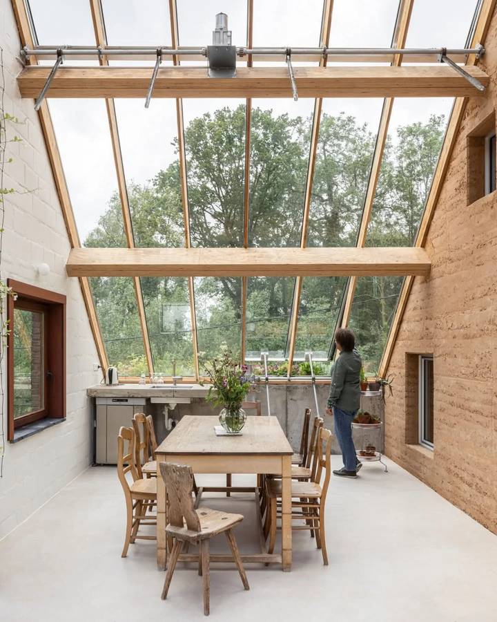 A dining room under a glazed lean-to roof, with a rammed earth wall on one side and a long timber table.