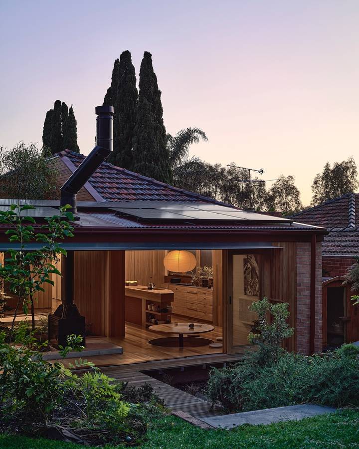 A brick house at dusk with a timber lined room open to a deck, seen across a planted garden.