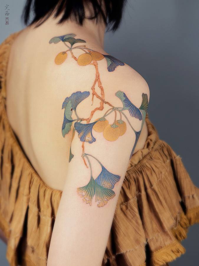 A tattoo of ginkgo leaves and orange fruit on a branch, running across a shoulder and upper arm.