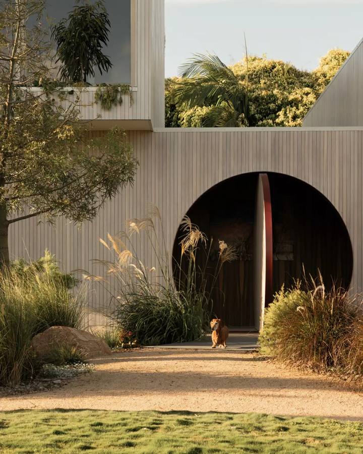 A timber battened house with a large circular opening at its entry, with grasses and a gravel path in front.