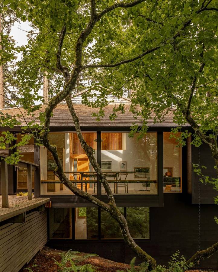 A dark timber midcentury house among maples, with full height glazing onto a dining room.
