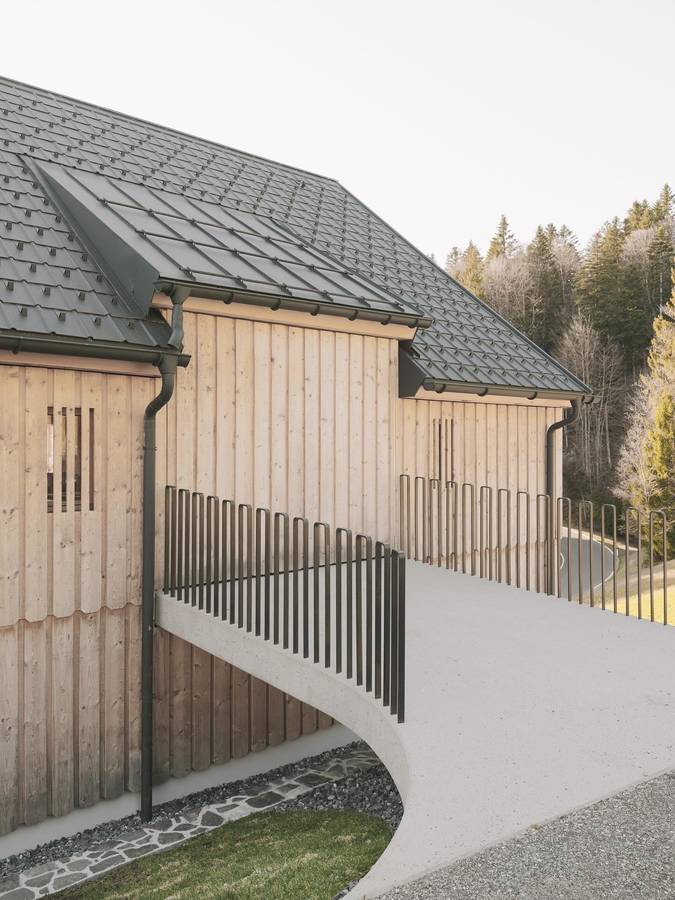 A timber clad alpine house with a dark shingled roof and a black slatted balustrade curving along a ramp.