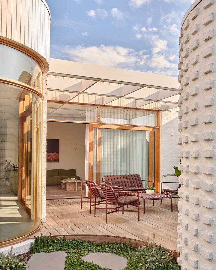 A timber deck between a curved white clad wall and a textured brick drum, with red steel outdoor chairs.