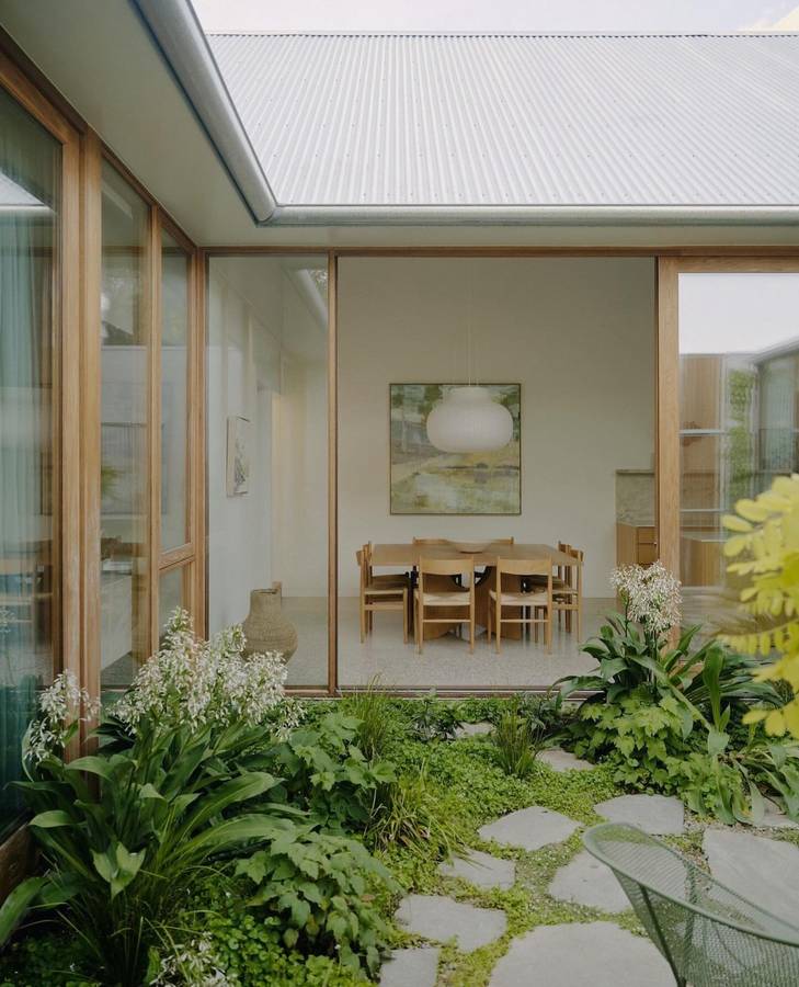 A planted courtyard under a corrugated roof, looking through glazing into a dining room.