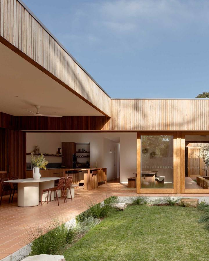 A timber clad courtyard house with terracotta paving, open to a kitchen and dining area beside a lawn.