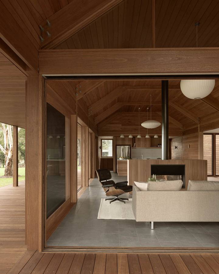 A timber lined room with a gabled ceiling seen from a verandah, with a kitchen and living space beyond.
