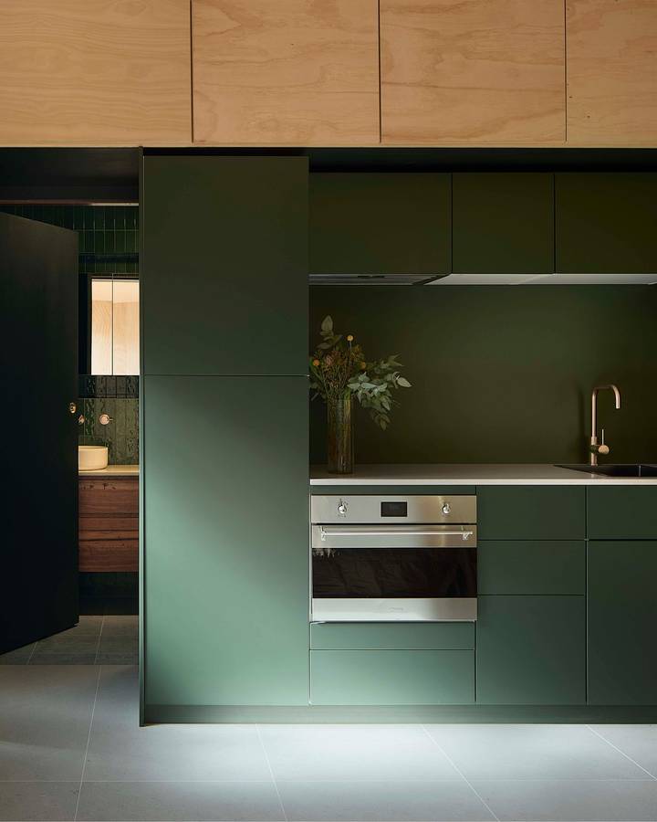 A kitchen in deep green cabinetry with plywood overhead cupboards and a brass tap.