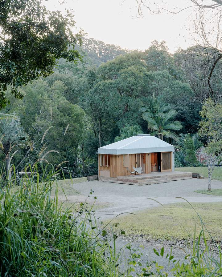 A small pale timber cabin with a hipped metal roof standing in a clearing above a gully of tree ferns and eucalypts.