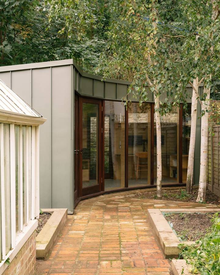 A pale metal clad garden room with a curved glazed end, seen along a brick path past a greenhouse and birch trunks.