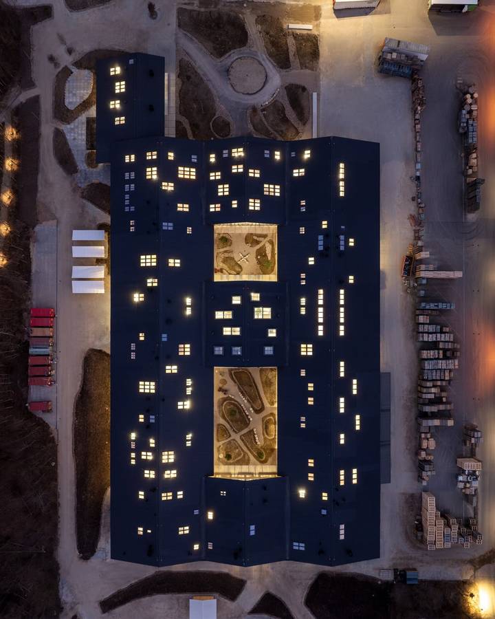 An overhead view at dusk of a long dark building with lit windows and two planted courtyards, surrounded by stacked timber.