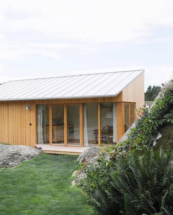 A small timber cabin with a folded metal roof and full height glazing, standing on a lawn beside a granite outcrop.