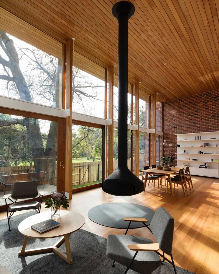 A living and dining room under a timber lined raked ceiling, with a suspended black fireplace and a wall of glazing to parkland.