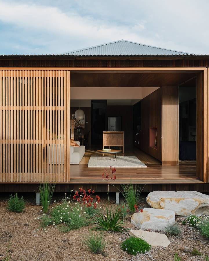 A timber battened screen slid back from an open living room under a corrugated roof, with a garden of rocks and grasses in front.