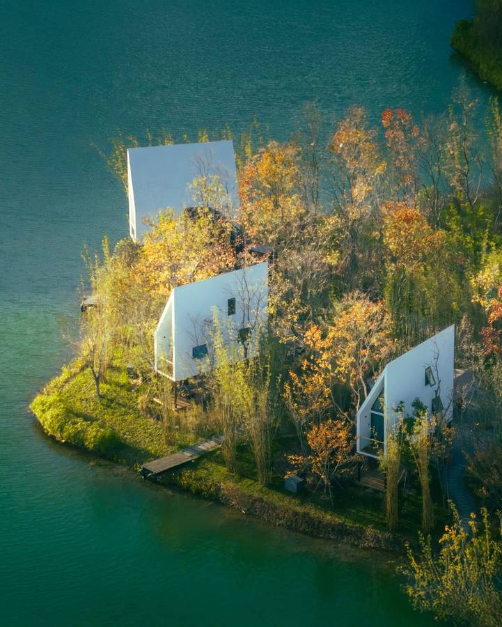 An aerial view of a wooded islet in green water, with narrow white cabins among autumn trees and a jetty at the shore.