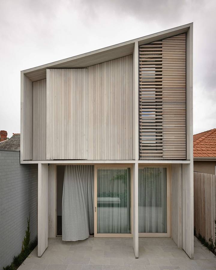 A pale timber clad upper floor with a battened screen, above a curtained glazed ground floor and a paved terrace.