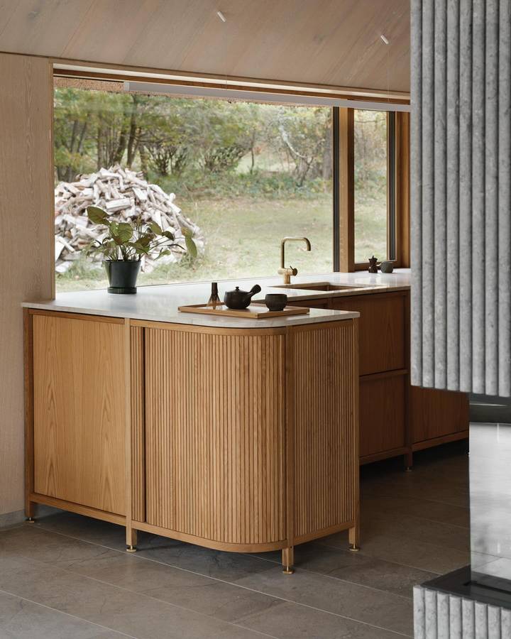 An oak kitchen with a fluted curved island and a stone benchtop, and a window over the sink looking out to a woodpile and trees.
