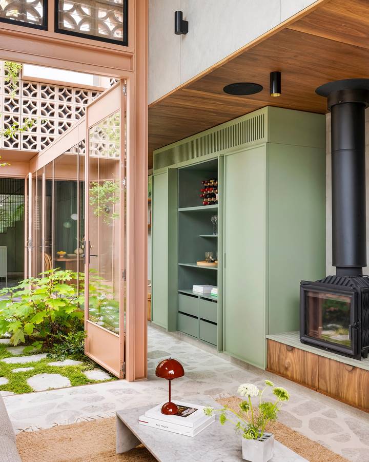 A living space with a green painted cabinet wall and a black wood heater, opening through glazed doors to a courtyard with stepping stones.