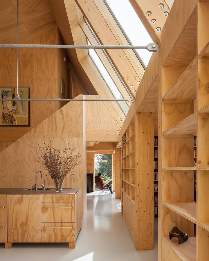 A plywood lined interior under a steeply pitched glazed roof, with a kitchen bench, tall shelving and a long room beyond.