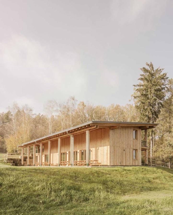 A larch clad building with a long mono pitched roof standing on a meadow at a woodland edge, with picnic tables along its front.