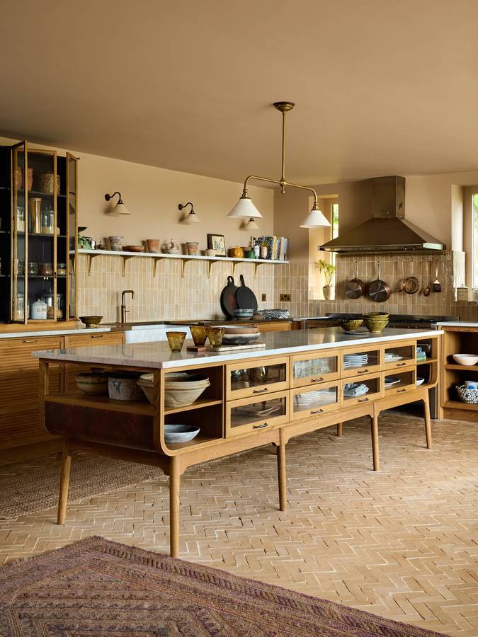 A kitchen with a freestanding oak and walnut island on turned legs, open shelves, a range under a canopy and a herringbone brick floor.