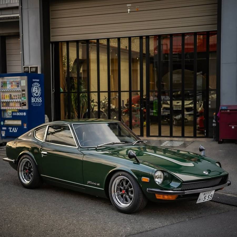 A dark green Datsun 240Z parked outside a glazed workshop, with a vending machine beside the door.