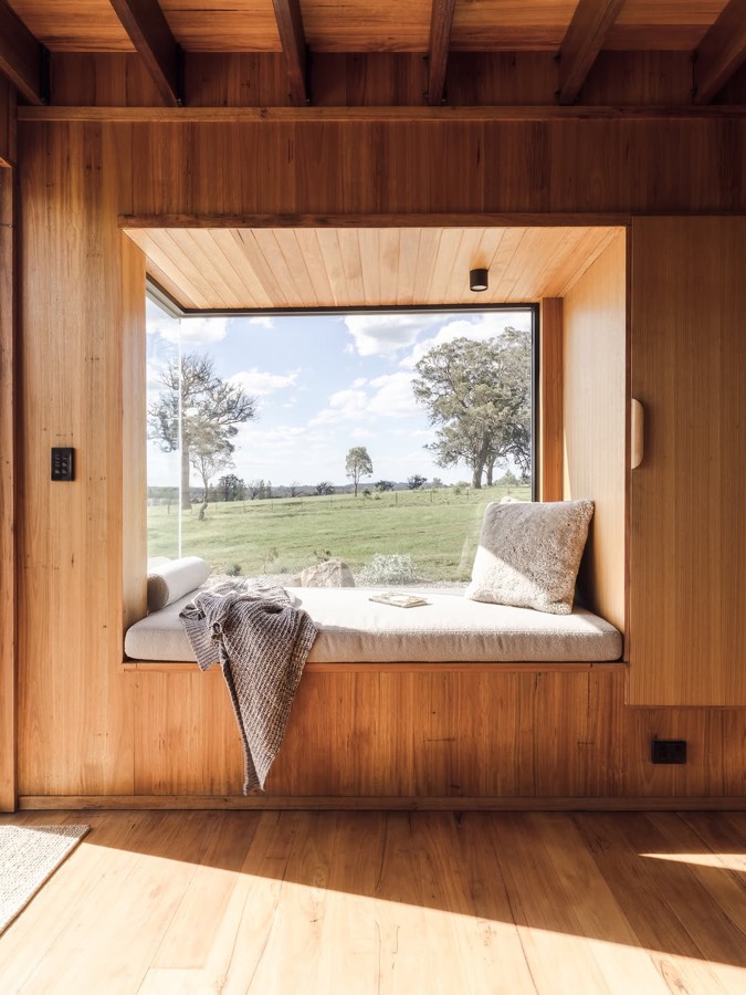 A timber lined window seat with a cushion and a blanket, looking out to a paddock and trees.