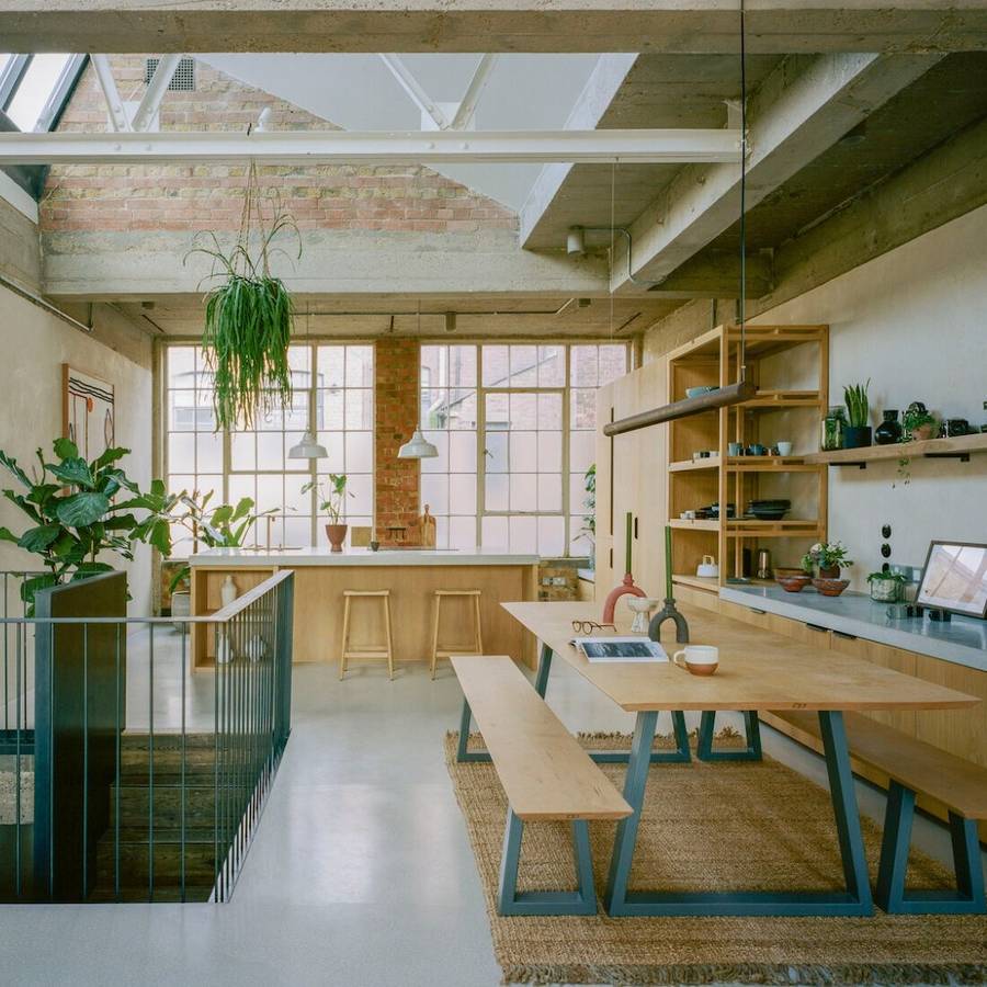 A converted factory interior under a north light roof, with a long table on trestle legs, open shelving and a kitchen beyond.