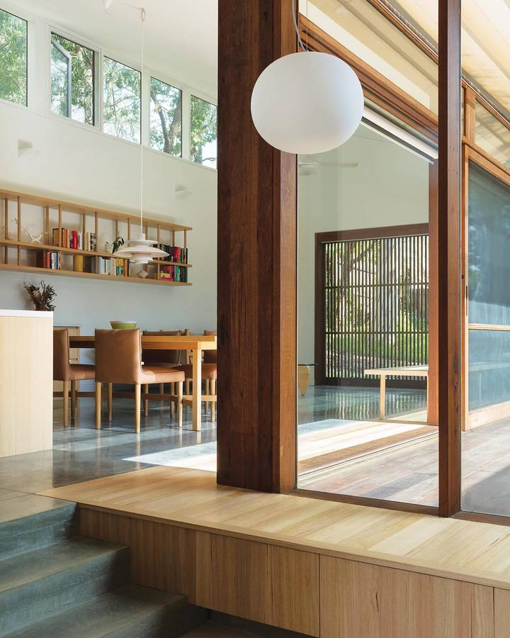 A living space with a full height glazed wall and a timber post, a white globe pendant over a dining table and a bookshelf along the wall.