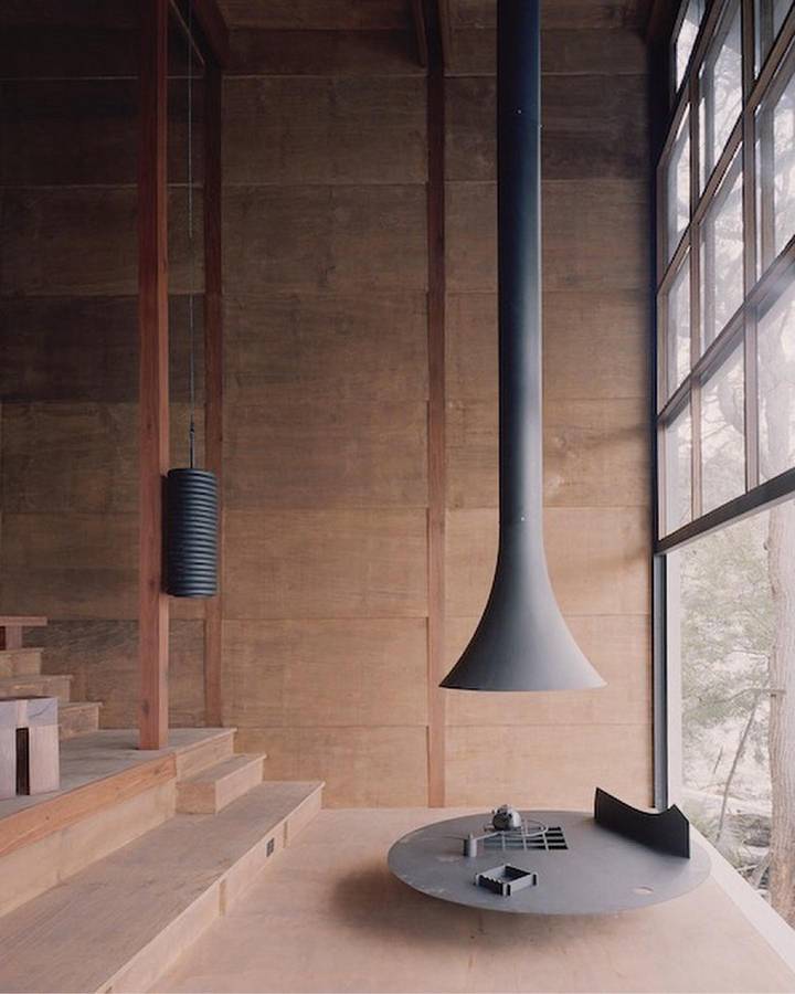 A tall room lined with wide timber boards, with a black flue widening into a hood over a round steel hearth beside a steel framed window.
