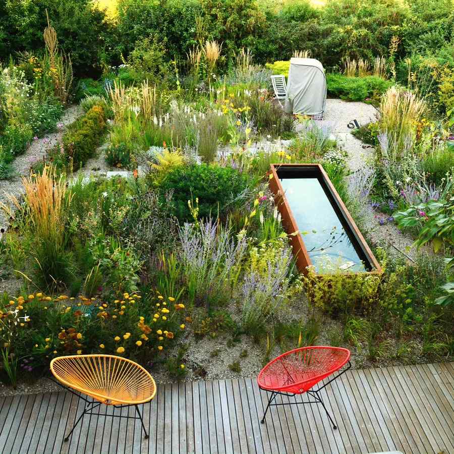 A sloping gravel garden of grasses and perennials seen from a timber deck, with a steel edged water basin set into the slope and two chairs on the boards.