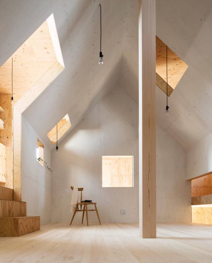 A white interior with a gabled inner form cut by angled openings, a plywood stair and two pendant bulbs.