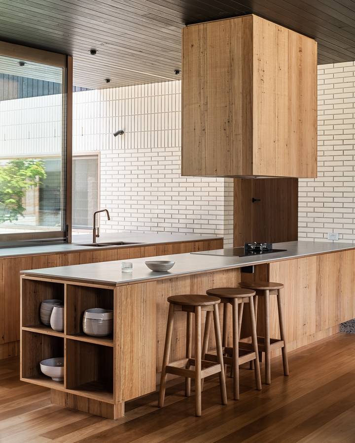 A kitchen with a timber island and a boxy timber rangehood against white glazed brick walls, with timber stools at the bench.