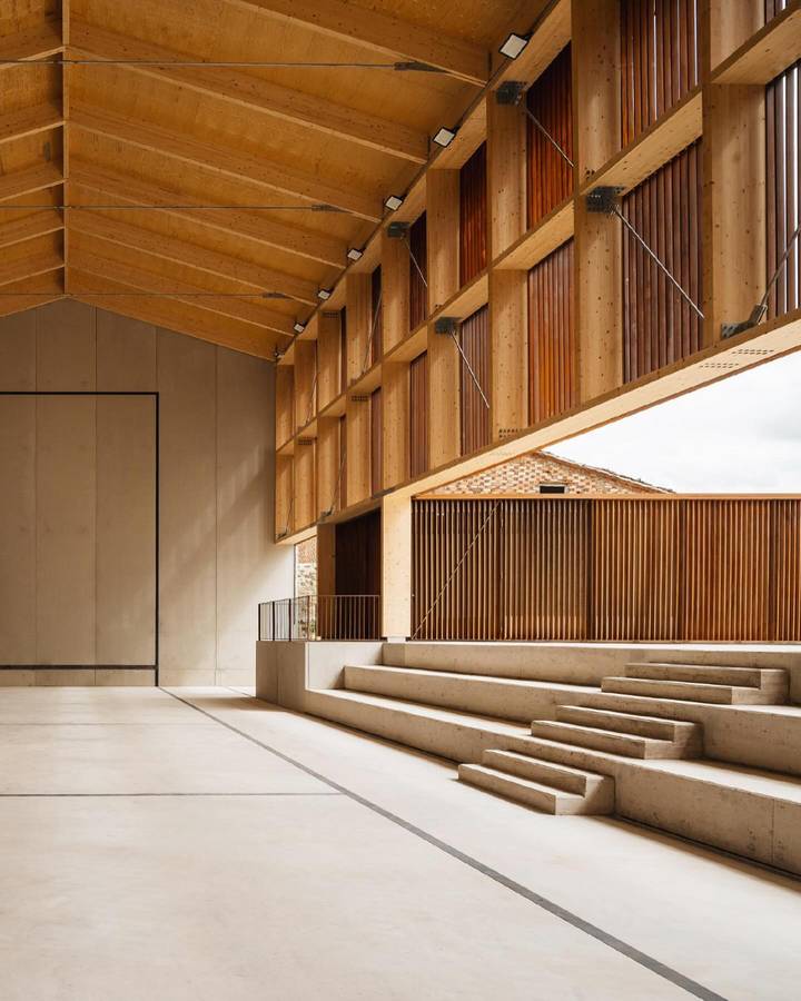 The inside of a pelota court under a timber roof structure, with concrete terracing and a slatted weathering steel screen at one end.