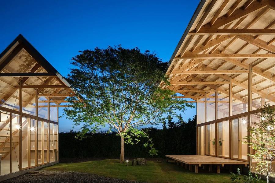 Two timber framed gabled pavilions facing each other across a lawn at dusk, with a lit tree between them.