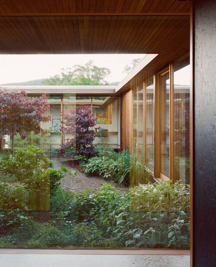 A planted courtyard seen through timber framed glazing under a timber lined soffit, with rooms on the far side and hills beyond.