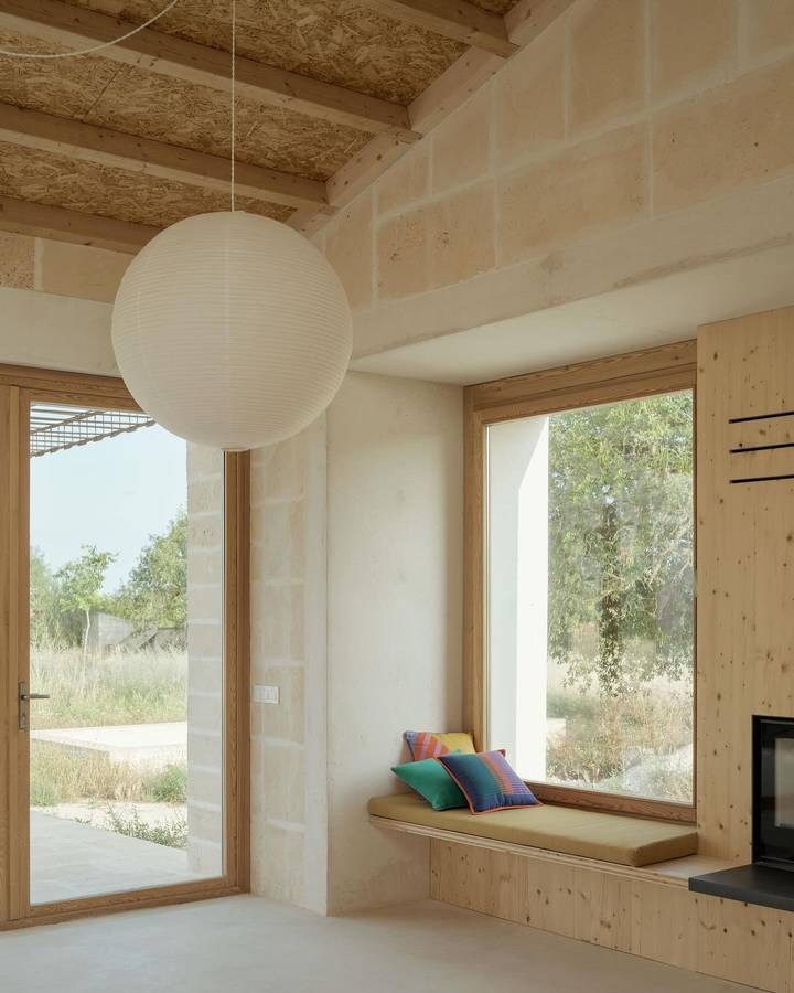 A room under a timber and reed ceiling on beams, with pale stone walls, a paper globe pendant and a timber window seat beside a wood heater.