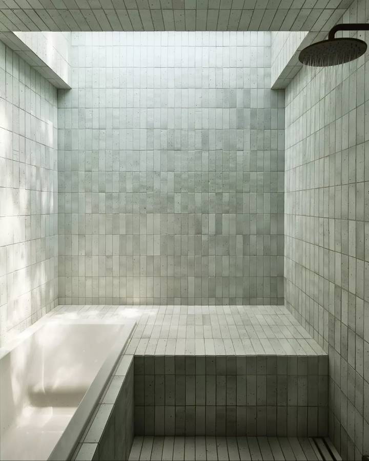 A bathroom lined in pale green square tiles, with a rain shower over a tiled bench and a bath at the near end.