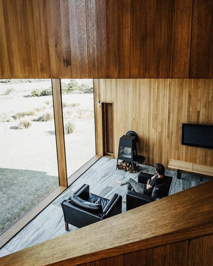 A timber lined room seen from above, with a wood heater, a low black sofa and a person reading beside full height glazing to heath.