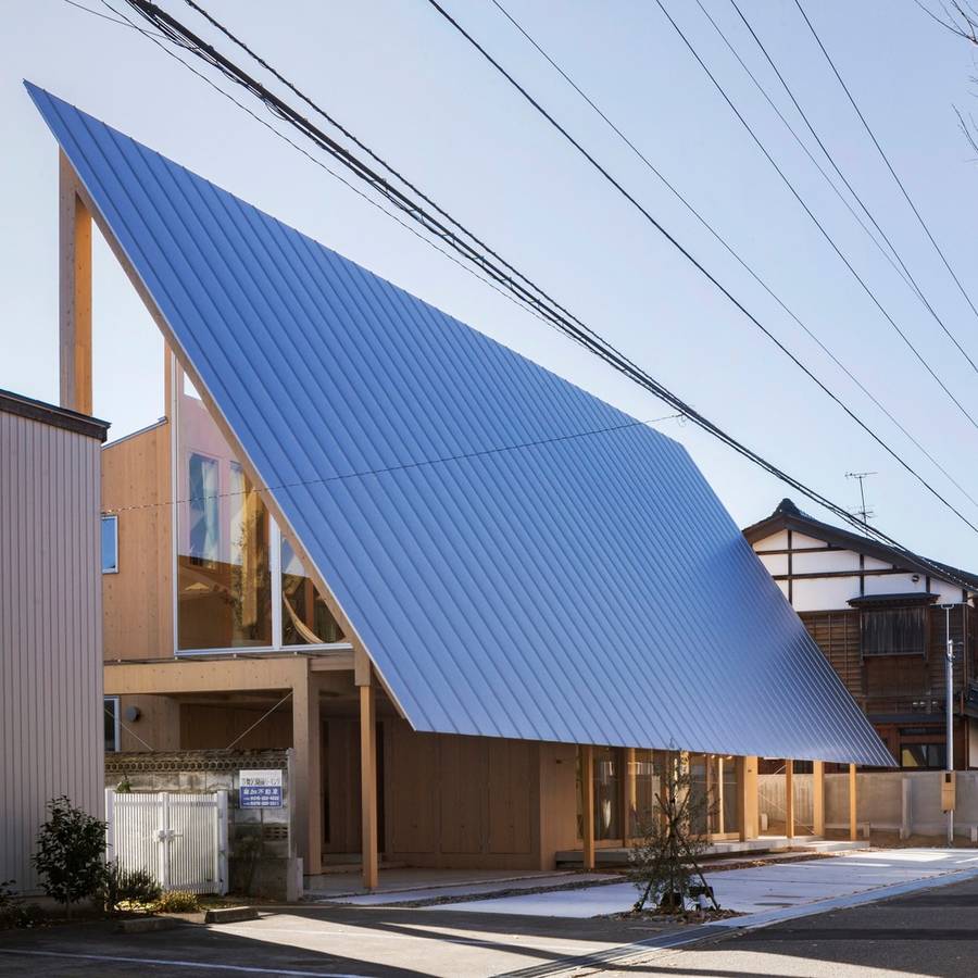 A house with a very large pitched metal roof sweeping down towards the street on timber posts, between neighbouring houses.