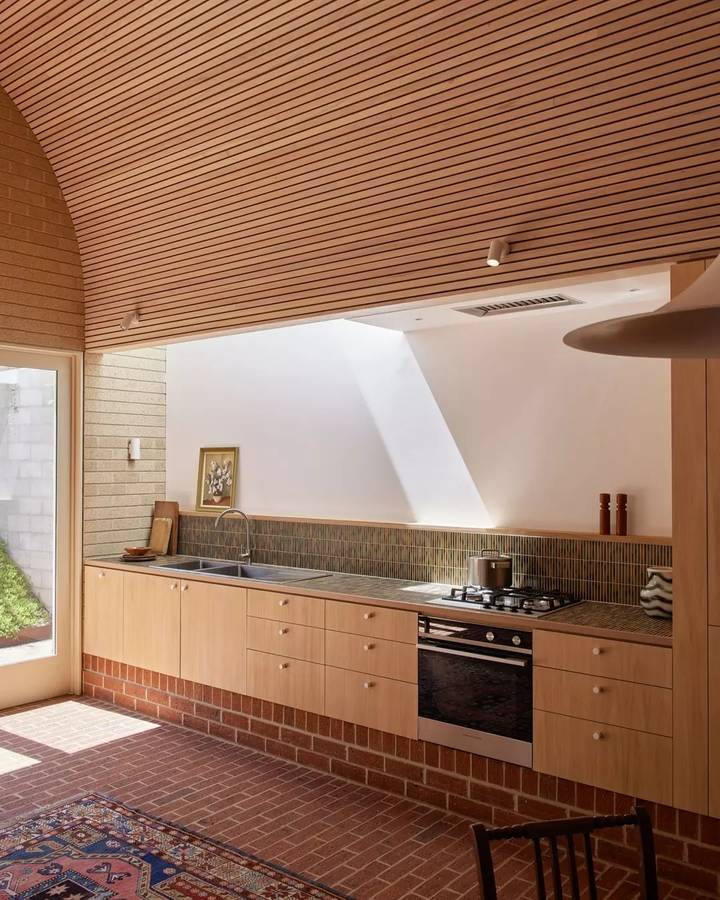 A kitchen under a curved timber battened ceiling, with pale timber cabinets, a green tiled splashback and a rug on a brick floor.