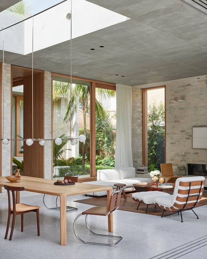 A living and dining room with a raw concrete ceiling and a long skylight, timber framed glazing to palms and a pale brick wall.
