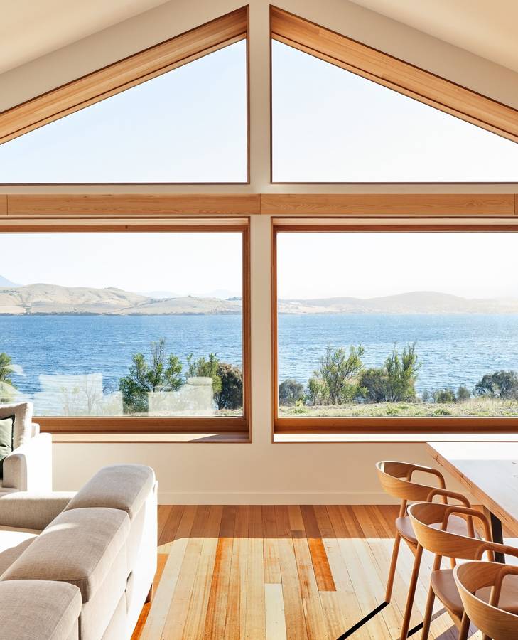 A room with a gabled timber framed window looking out to a bay and hills, with a pale sofa and dining chairs on a timber floor.
