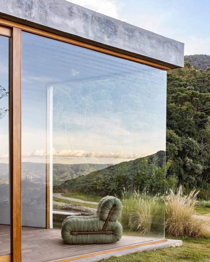 A glazed corner of a whitewashed earth walled house looking out over a forested valley, with a round cushioned seat on a brick terrace.