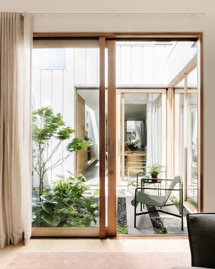 A planted courtyard seen through timber framed sliding doors, with white board walls and a green wire chair.