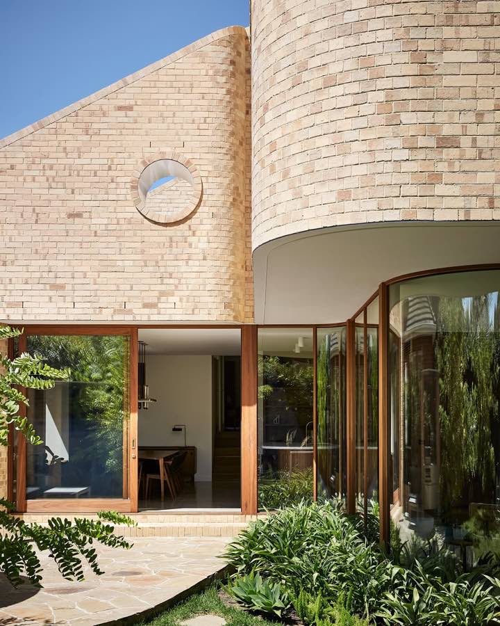 A curved brick wall with a round window beside timber framed glass doors, with planting in front.
