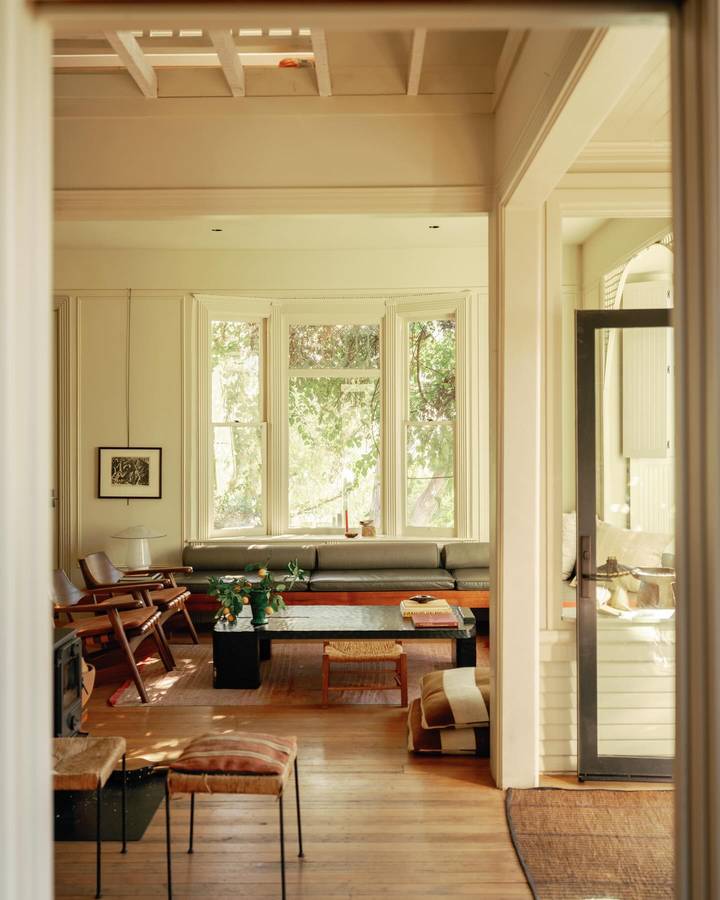 A cream painted timber room seen through a doorway, with a green banquette under casement windows and leather stools on a timber floor.