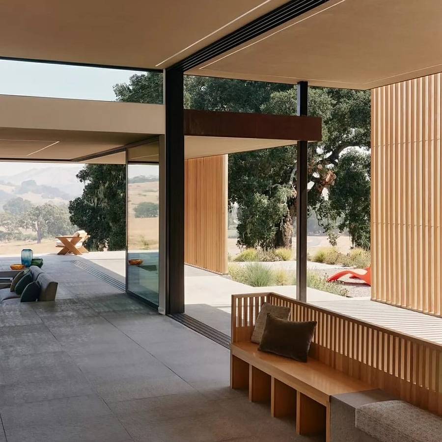 A covered outdoor room with a timber battened wall and a built in bench, sliding glass to a living room, with oaks and dry hills beyond.