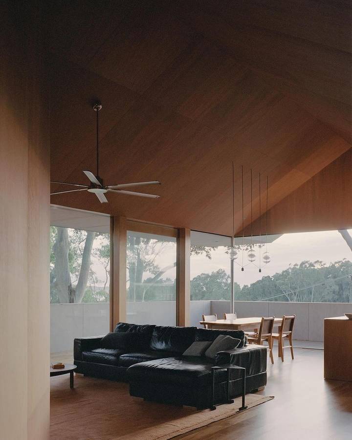 A living room under a dark timber raked ceiling with a ceiling fan, a black leather sofa and full height glazing to bush.